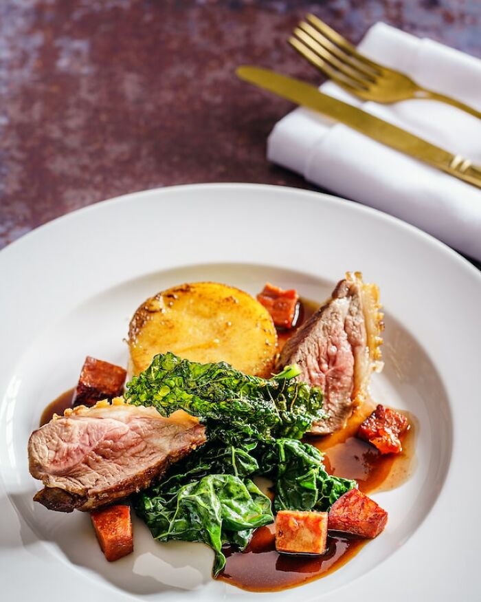 Plated gourmet meal featuring seared meat, sautéed greens, roasted root vegetables, and a rich sauce on a white plate.
