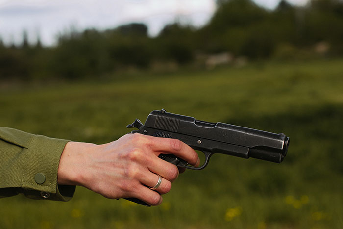 Hand holding a black handgun outdoors with blurred green nature background, unrelated to texting games or phone conversation.