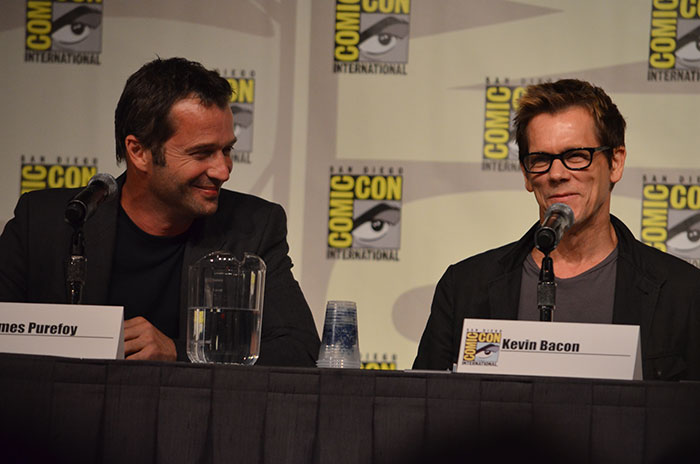 Two men speaking at a panel discussion with microphones and Comic-Con International banners in the background, engaging in texting games.