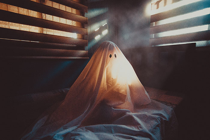 Person dressed as a ghost with a white sheet and cut-out eyes sitting in a dimly lit room with blinds.