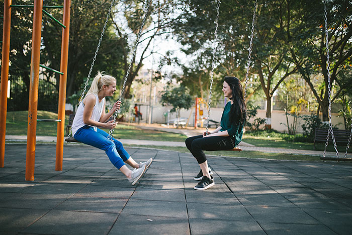 Two young women on swings in a park, smiling and engaged in a lively texting games conversation outside.