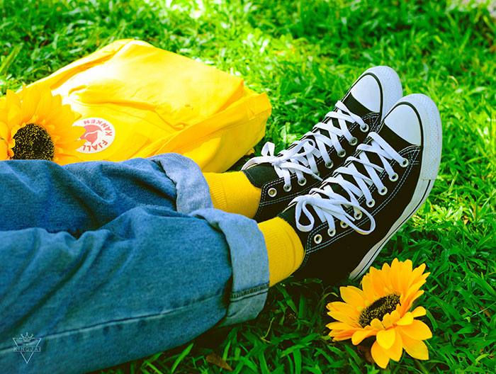 Person wearing black sneakers and yellow socks resting on grass with a yellow backpack and sunflowers nearby for texting games.