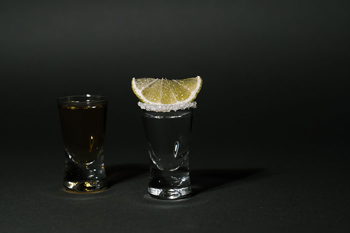 Two shot glasses with clear and dark liquid, one rimmed with salt and topped with a lemon wedge, on a dark surface.