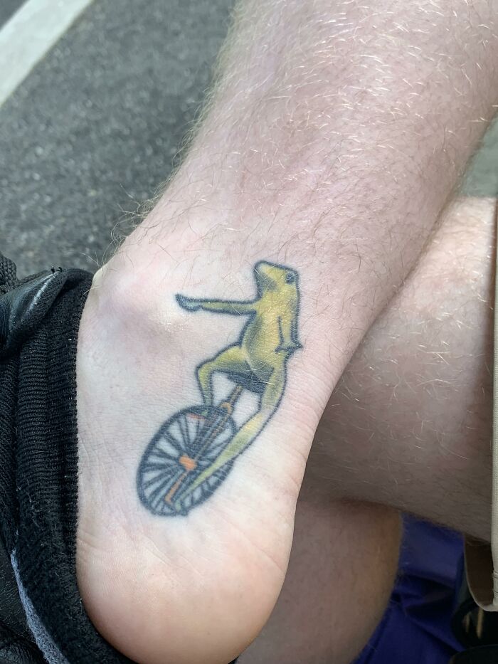 Tattoo of a yellow unicycling frog on a person's ankle, showcasing a cringe-worthy design.