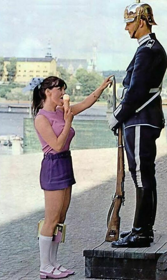 1970s. Girl Licking Ice Cream And Playing With The Tip Of The Gun’s Bayonet To Elicit A Reaction From A Royal Guard Outside Stockholm Palace, Stockholm, Sweden