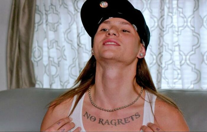 Person showing a misspelled "No Ragrets" tattoo on chest, wearing a hat and tank top, illustrating cringe-worthy tattoo.