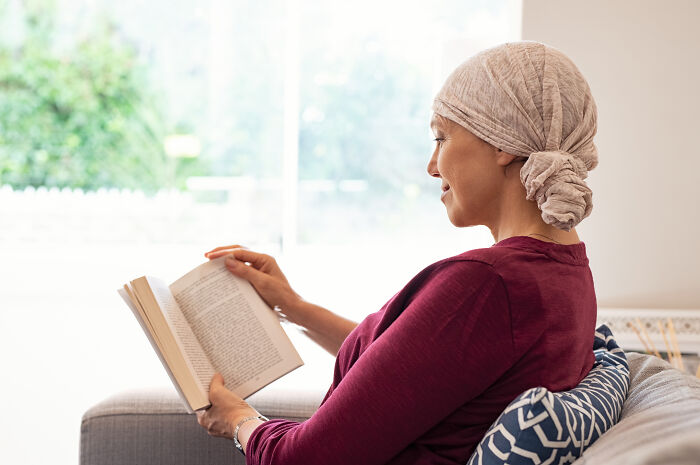 Person in a maroon top and headscarf reading a book, seated on a couch, with a bright window in the background.