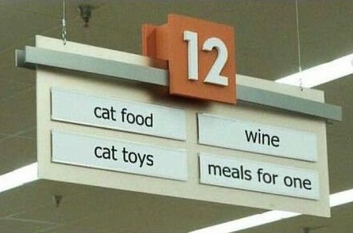 Grocery aisle sign showing cat food, cat toys, wine, and meals for one, a hilariously absurd store label.