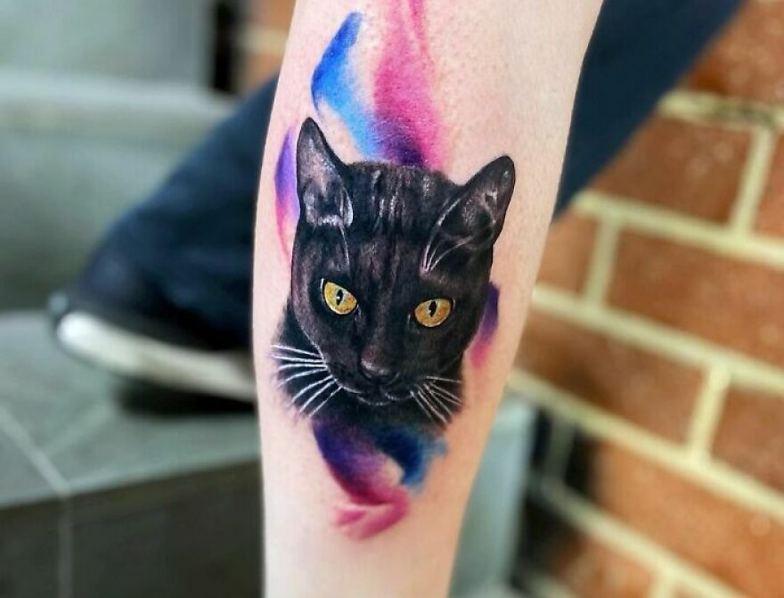 Watercolor tattoo of a black cat with vibrant pink and blue background on arm.