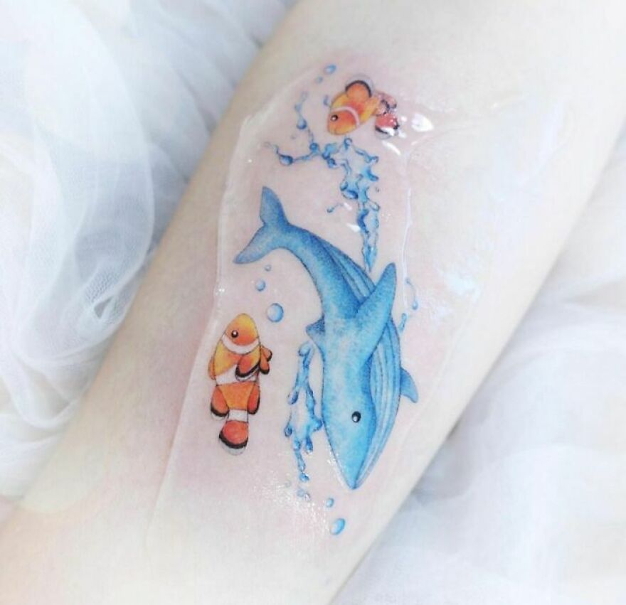 Watercolor tattoo of a blue whale and clownfish with splashes of water on skin.