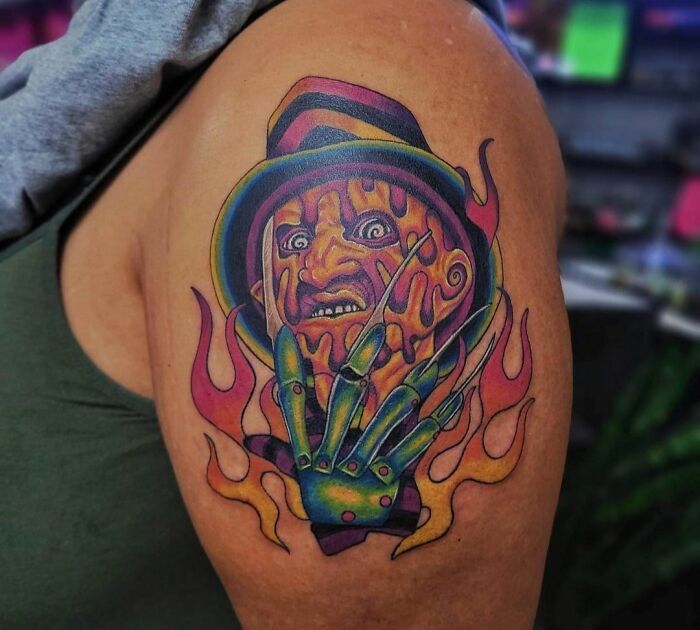 Colorful psychedelic tattoo featuring a surreal face with vivid flames on a person's arm.