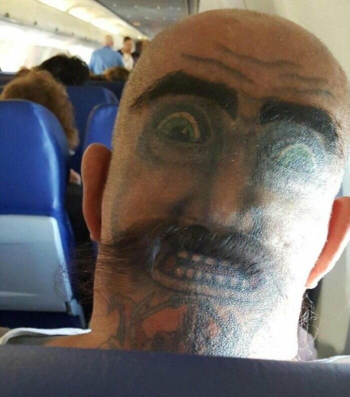 Funny face tattoo on back of head in an airplane seat, showcasing unique body art.