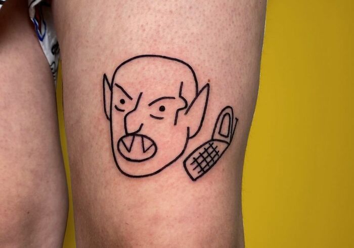 Funny tattoo of an angry person with pointy ears on a leg, featuring a simplistic design.