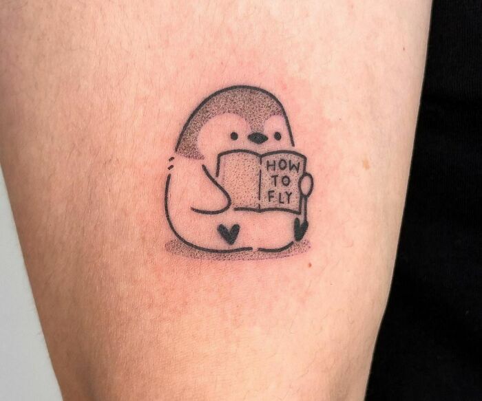 Funny tattoo of a fat penguin reading "How to Fly" on a person's arm.