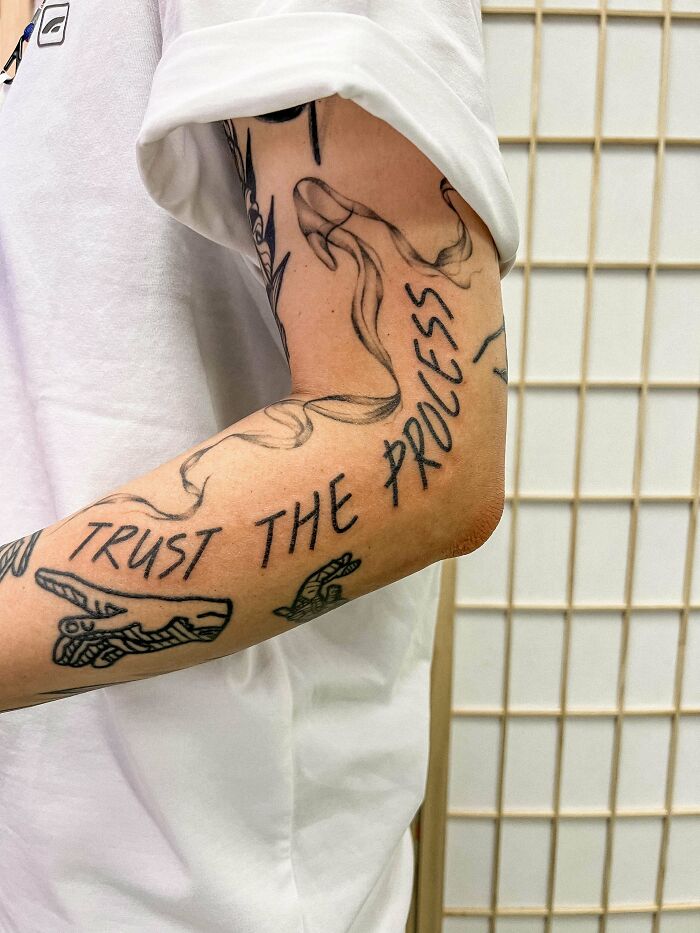 Arm with artistic word tattoo "Trust the Process" and sketch-style designs.