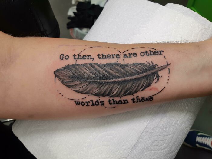 Feather tattoo with the phrase "Go then, there are other worlds than these" on an arm.