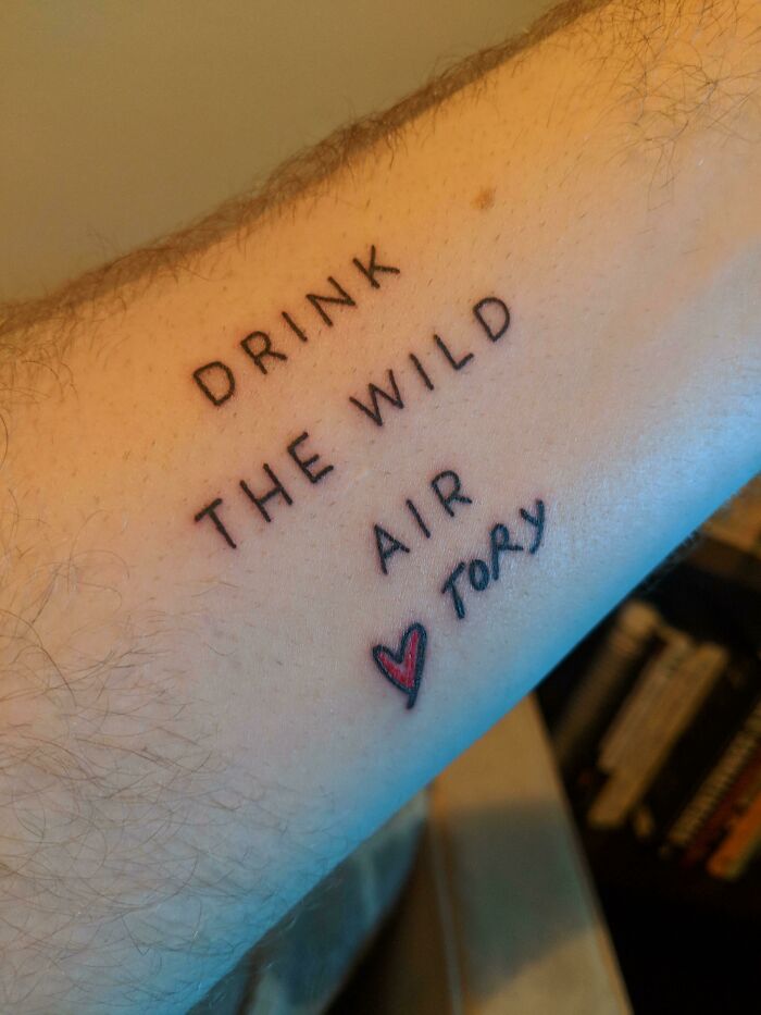 Word tattoo reading "Drink the wild air" with a red heart on an arm.