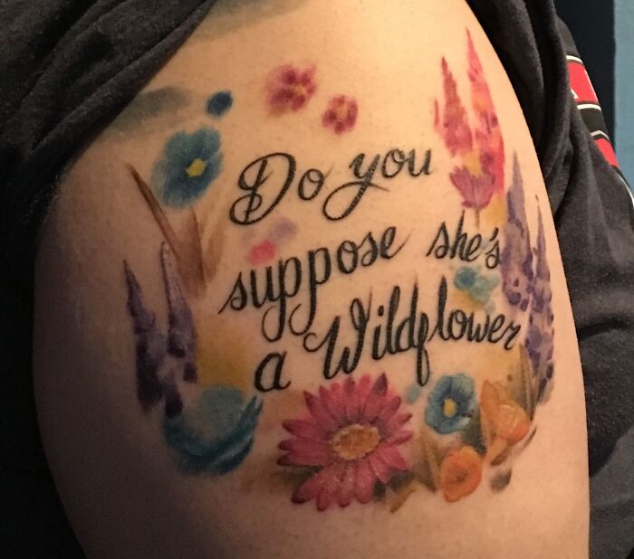 "Script tattoo with 'Do you suppose she's a Wildflower' and colorful floral designs on an arm."