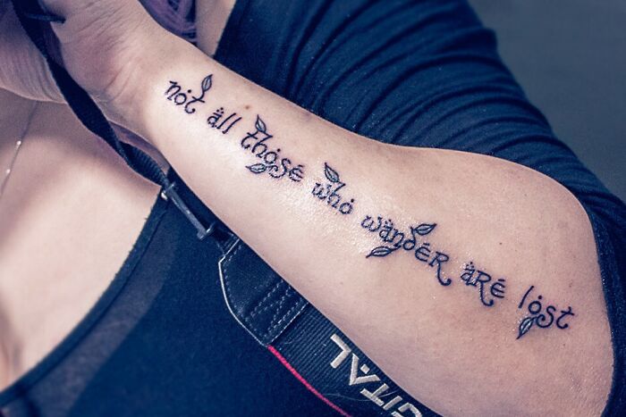 Word tattoo with the phrase "not all those who wander are lost," featuring ornate text on a person's forearm.
