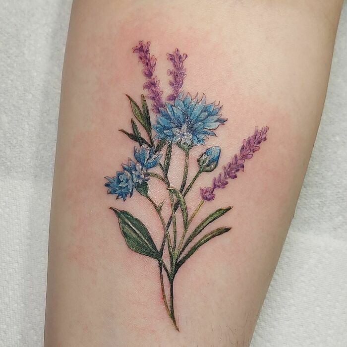 Nature tattoo featuring blue and purple flowers on skin, celebrating the beauty of Mother Earth.