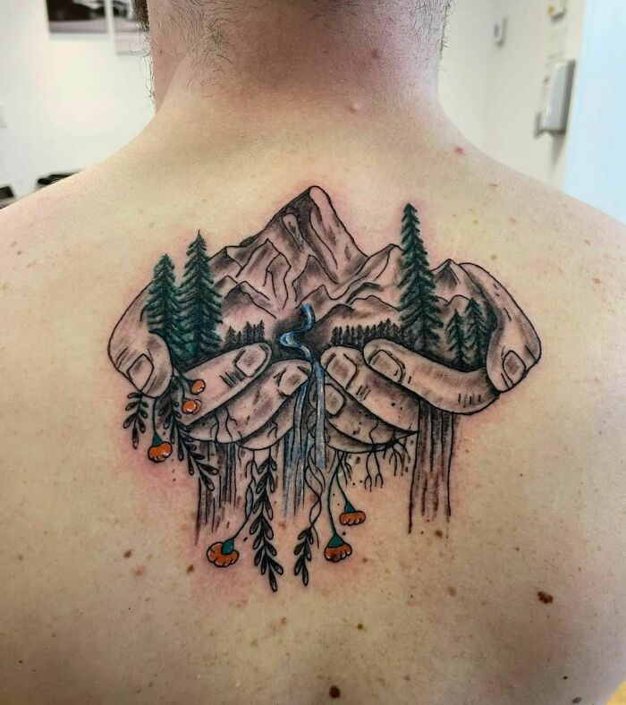 Nature tattoo featuring hands cradling mountains and trees on a person's back, symbolizing the human-earth bond.