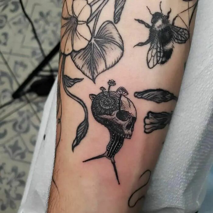 Nature tattoo on arm featuring a bee, flowers, leaves, and a mushroom-topped skull, symbolizing human-earth connection.