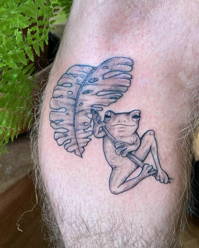 Nature tattoo featuring a frog holding a leaf on the leg, with a green plant in the background.