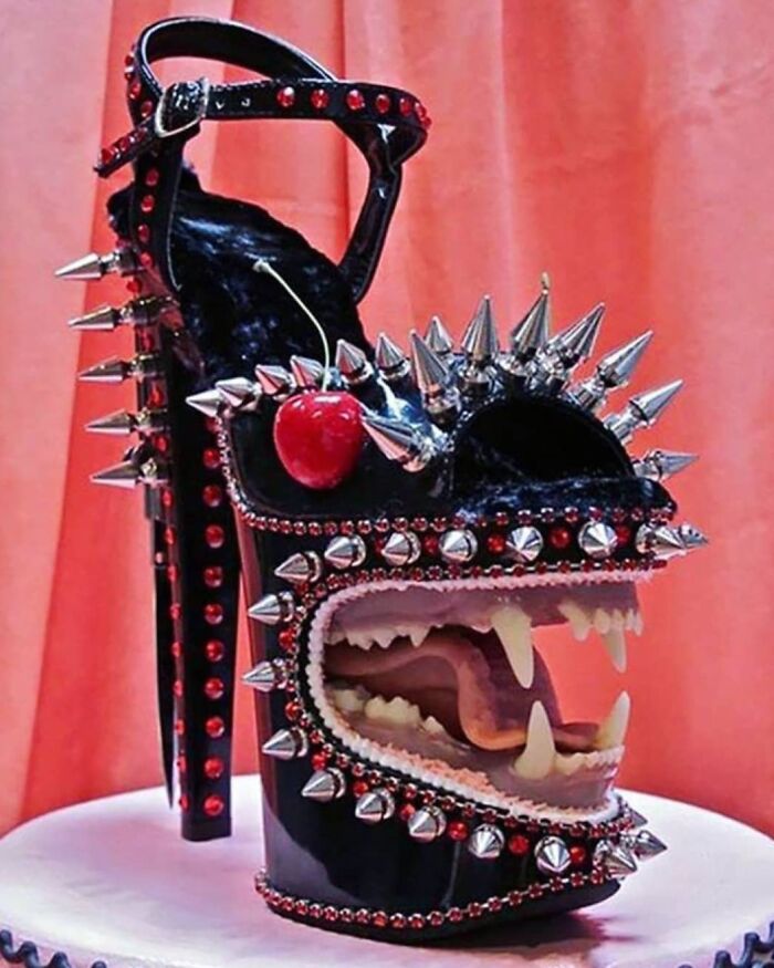 Weird high heel shoe with spikes, studs, and a cherry detail on a pink background.