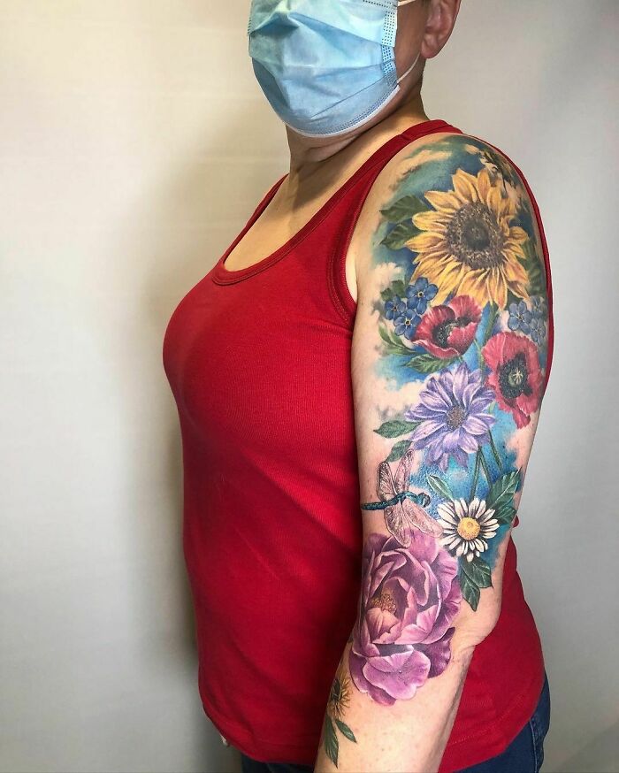 Person with nature tattoo featuring colorful flowers and a dragonfly on their arm, wearing a red tank top and mask.