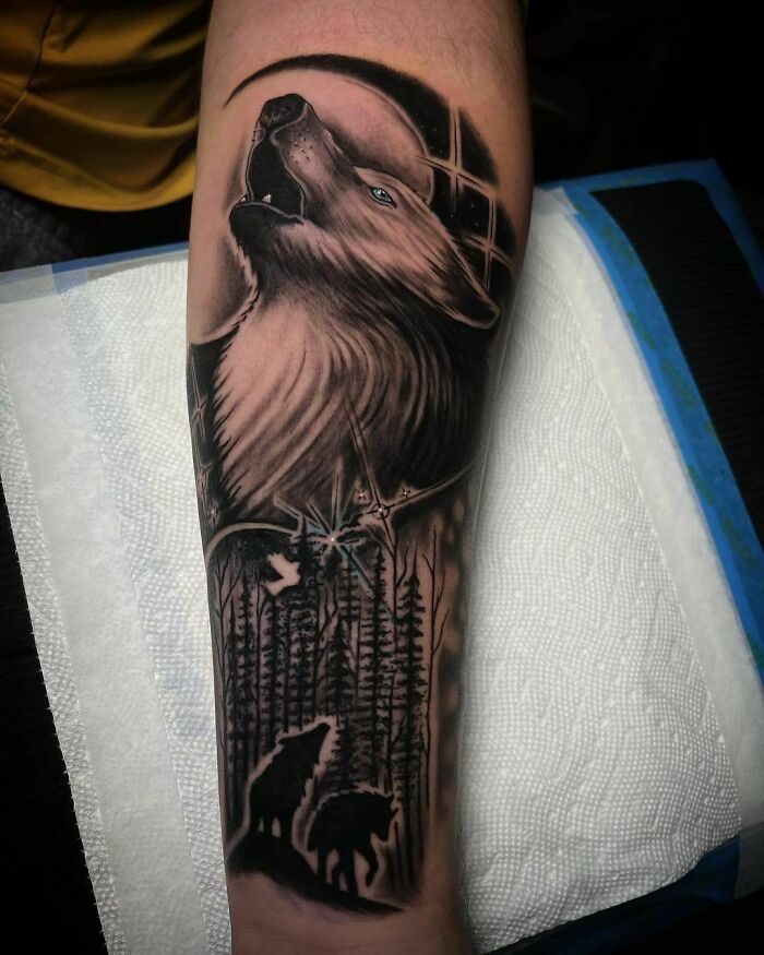 Nature tattoo depicting a howling wolf and forest scene on an arm.