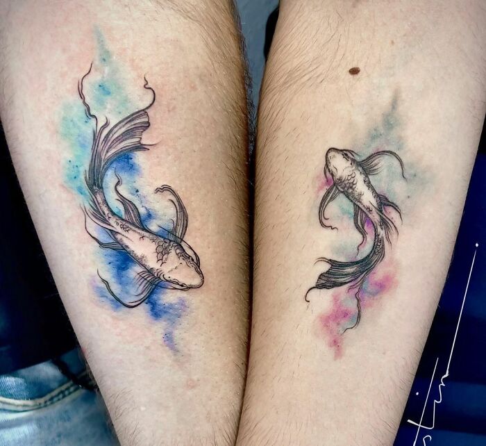 Watercolor tattoos of two fish with vibrant blue and pink hues on forearms, showcasing ethereal artistry.