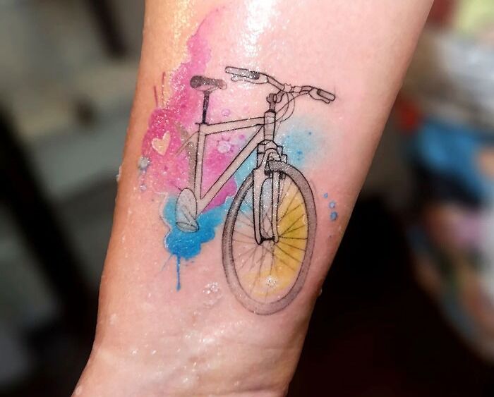 Watercolor tattoo of a bicycle with colorful, ethereal splashes of pink, blue, and yellow on skin.