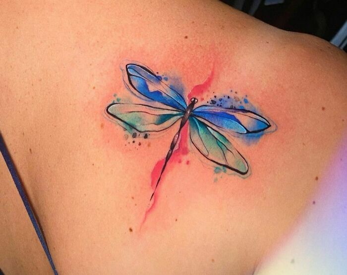 Watercolor tattoo of a dragonfly on shoulder, featuring ethereal blue and green hues with a soft pink background.