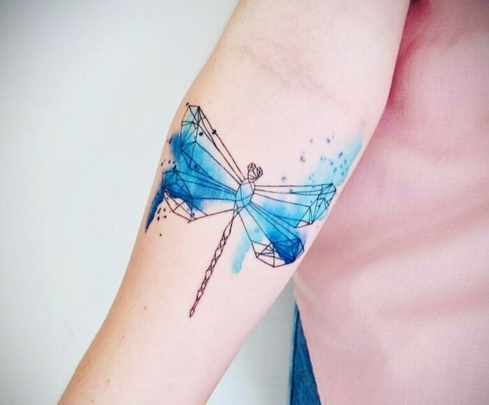 Watercolor tattoo of a blue and black geometric dragonfly on an arm.