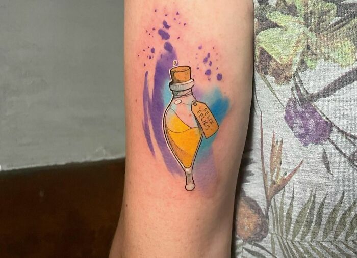 Watercolor tattoo of a potion bottle labeled "Felix Felicis" with purple and blue splashes on an arm.