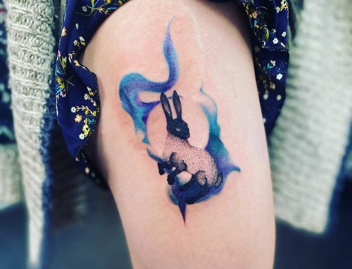 Watercolor tattoo of a rabbit with blue and purple hues on a person's leg, showcasing ethereal artistry.