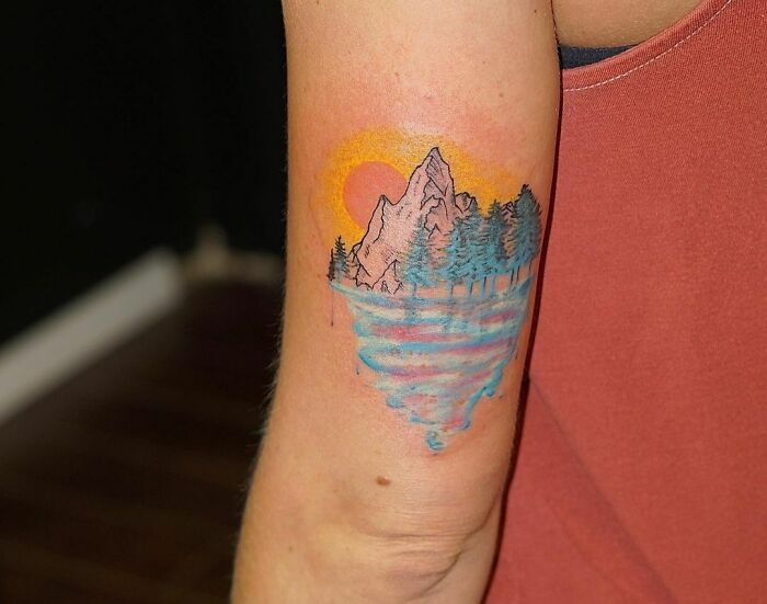 Watercolor tattoo of a mountain landscape with trees and a sunset on an arm.