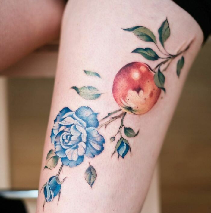 Watercolor tattoo of a blue rose and red apple on a person's leg, showcasing ethereal floral design.