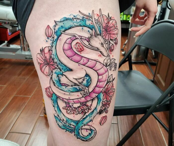 Watercolor tattoo of a dragon and flowers on a person's thigh, blending vibrant pink and blue hues.