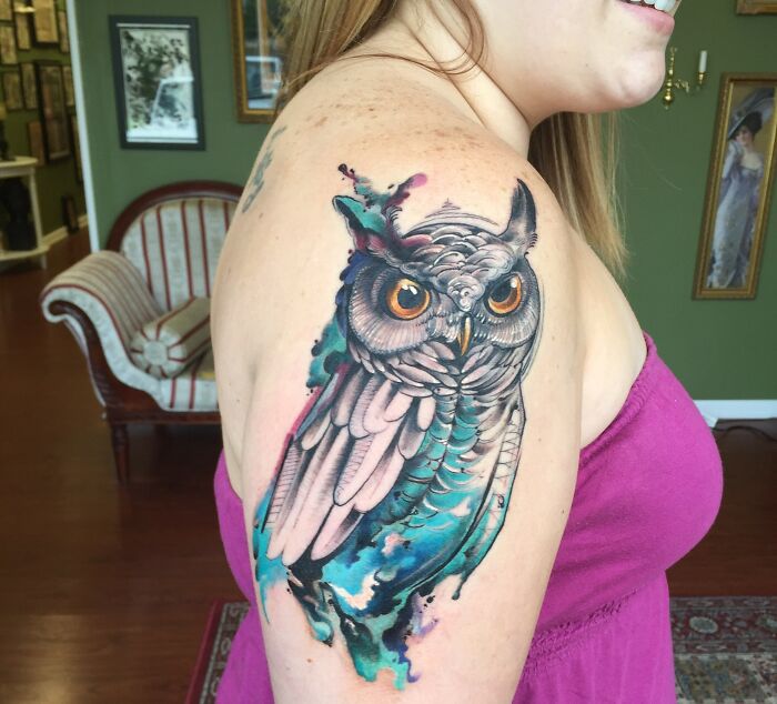 Woman with ethereal watercolor tattoo of an owl on her upper arm, indoors.