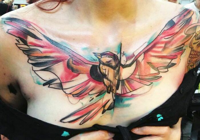Colorful bird tattoo art on woman's chest, featuring vibrant watercolor wings.