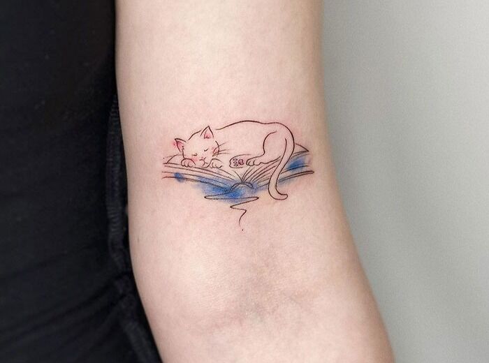 Minimalist animal tattoo of a sleeping cat on an arm, accented with subtle blue shading.