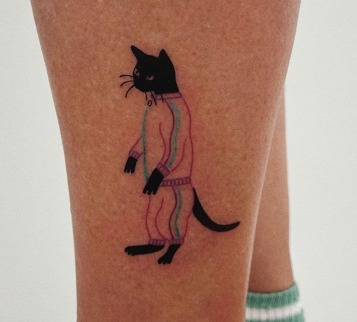 Black cat in a tracksuit tattoo on a person's leg, showcasing unique animal tattoo design.