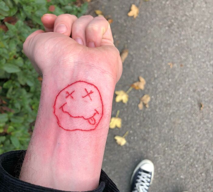 Funny tattoo of a red outline smiley face on a wrist, depicting a playful expression with crossed-out eyes and a tongue out.