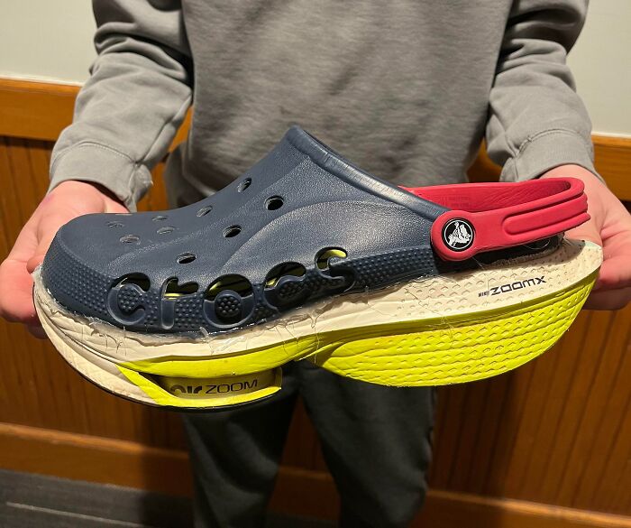 A person holding a weird shoe design featuring a large Croc with stacked sneaker soles.