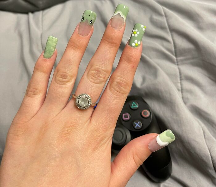 Hand with green nail art ideas featuring playful designs and patterns, holding a black gaming controller on a grey surface.