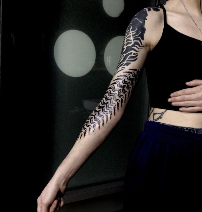 Arm with a geometric tattoo featuring intricate black patterns against a dark background.