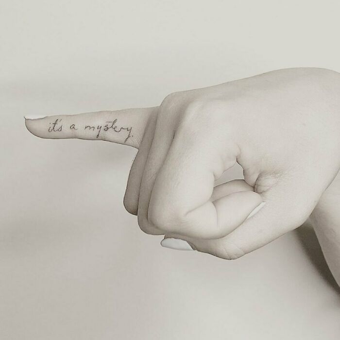 Close-up of a small celebrity tattoo on a finger reading "it's a mystery" in elegant script.