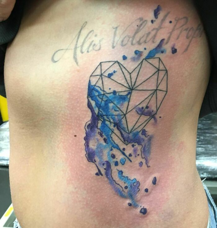 Geometric heart tattoo with blue watercolor accents and script text on skin.