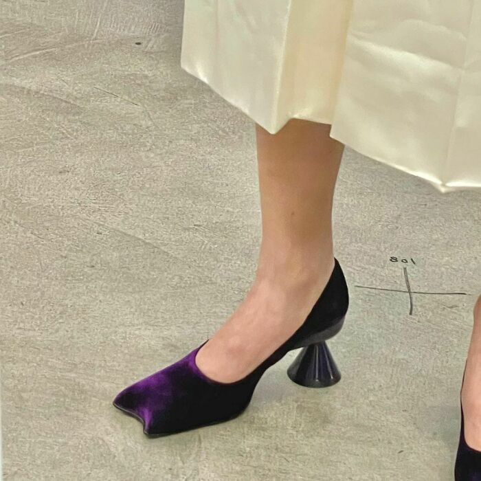 Unusual purple velvet shoes with unique conical heels on a person wearing a white skirt.